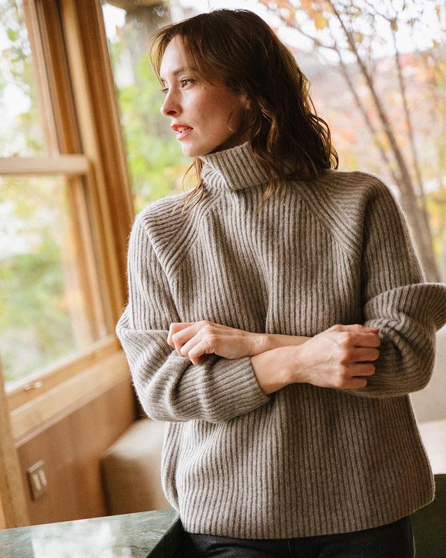 Woman in a natural grey ribbed yak turtleneck sweater standing by a wooden window frame