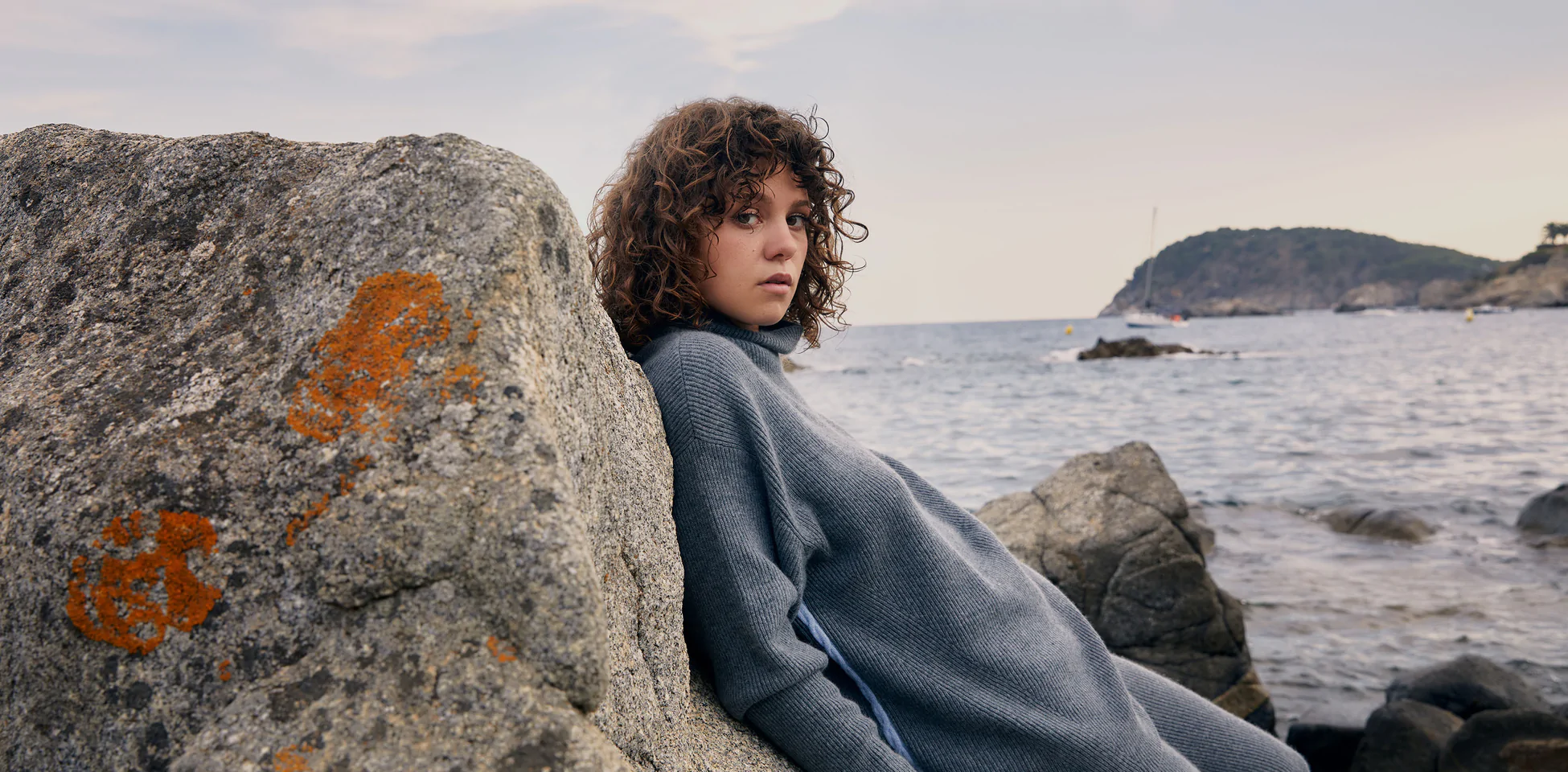 Model leaning against a coastal rock in an oversized grey ribbed knit, with the sea behind her
