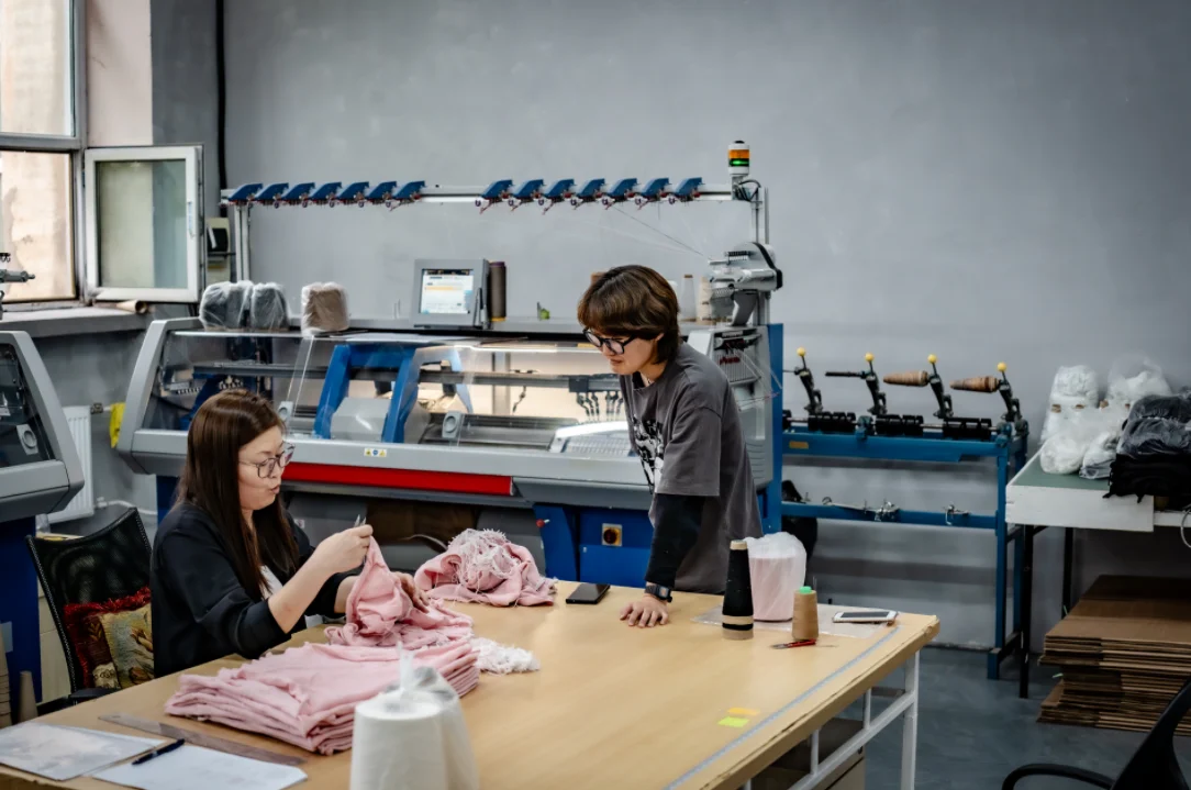 Selenge and Naraa from the HYPECHASE team working on a small-batch knitwear order beside a flat knitting machine in the atelier in Ulaanbaatar, Mongolia